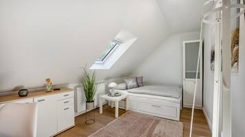 Une pièce compacte et bien éclairée, avec un plafond en pente, une lucarne, un lit, une petite table et un meuble blanc.