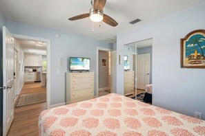 Ein Schlafzimmer mit Bett, Deckenventilator, Fernseher und einer Küche im Hintergrund.