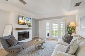 A living room with a fireplace, a television mounted above it, a sofa, a coffee table, and a door leading to a balcony. - Bay Breeze Bungalow (Anna Maria)