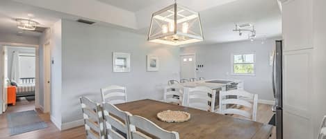Et spiseområde med et træbord og hvide stole, en hængende lampe, samt et køkken med hvide skabe.