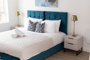 A neatly made bed with a blue headboard, white bedding, and patterned pillows. There are two bedside tables with lamps and a small drawer unit. - Modern 2-Bed Flat 70 at Sapphire Court, Birmingham (West Midlands)