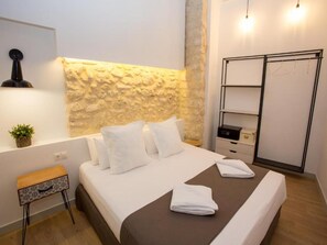 A modern hotel room with a stone accent wall, a neatly made bed with white linens, a wooden side table, and a built-in wardrobe. - Deluxe Apartment With Jacuzzy (Alicante)