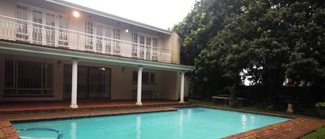 A rectangular swimming pool with a brick border, surrounded by grass and a building with a balcony.