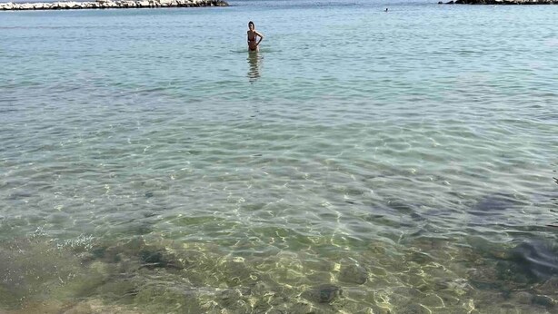 Une plage avec une eau claire et peu profonde, une personne debout dans l’eau et une côte rocheuse en arrière-plan.