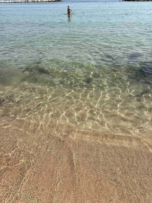 A beach with clear shallow water, a person standing in the water, and a rocky shoreline in the background. - Apartment by the Sea (Bari)