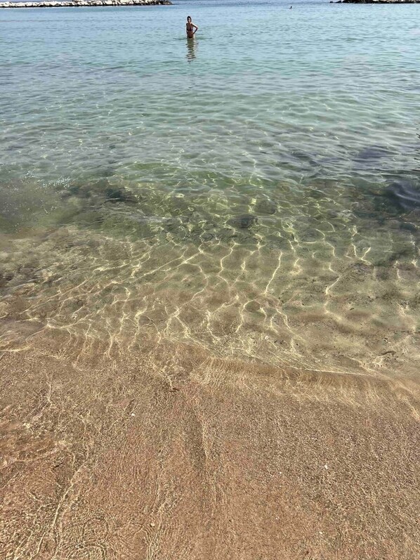 A beach with clear shallow water, a person standing in the water, and a rocky shoreline in the background. - Apartment by the Sea (Bari)