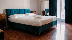 Un lit bien fait, avec une tête de lit turquoise et du linge de lit blanc, dans une pièce dotée d’un plancher ciré et d’une grande fenêtre agrémentée de rideaux.