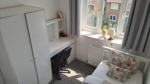 Een compact, net kamer met een bureau, een radiator, een raam met gordijnen en uitzicht op gebouwen.