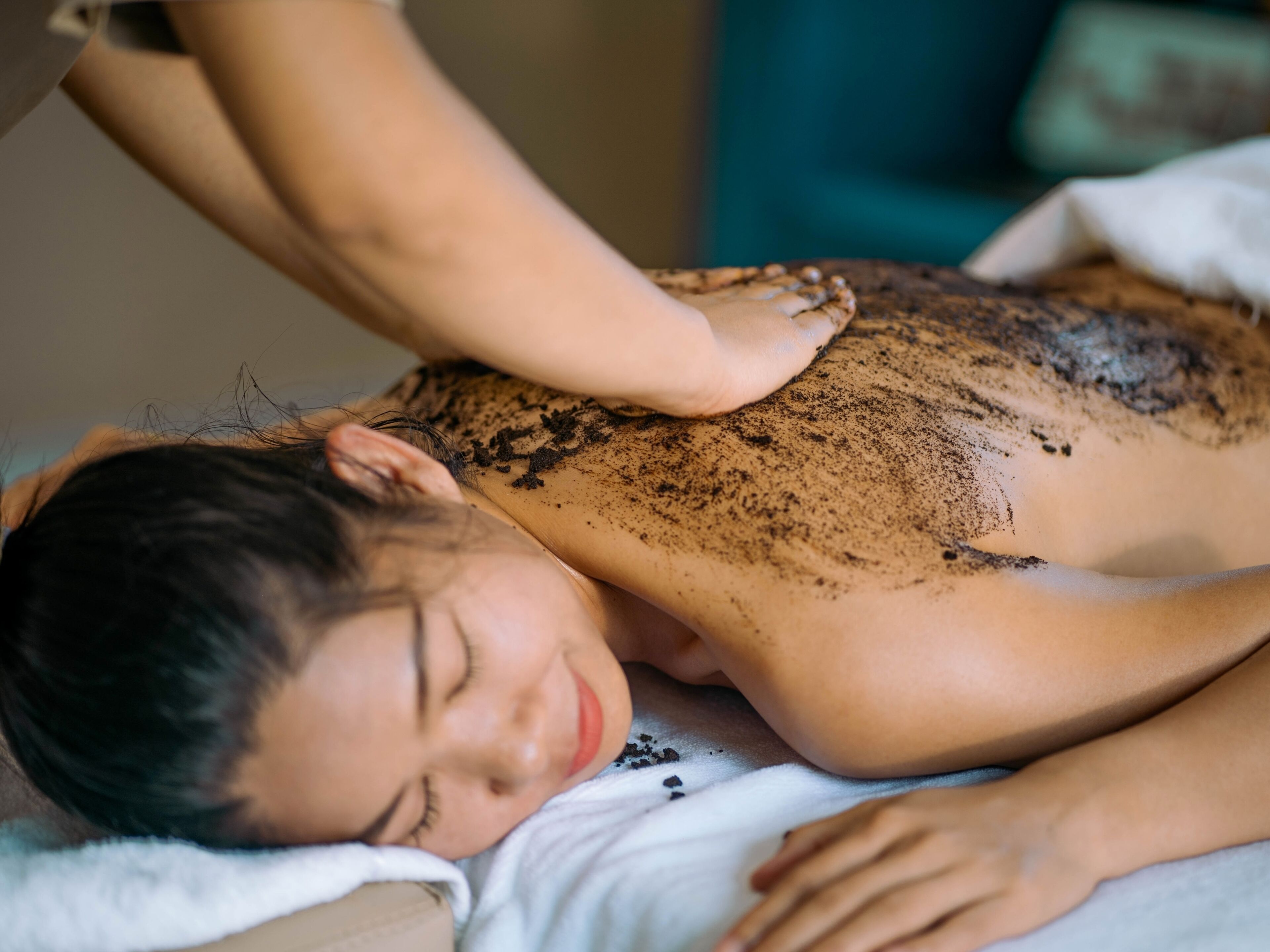 A person lying on a bed with a white sheet, receiving a back treatment with a brown substance.