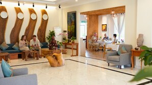 Un hall d’hôtel comprenant un comptoir d’accueil, un espace salon avec des canapés et des fauteuils, des éléments de décoration murale en bois, et un coin repas avec une table et des chaises.