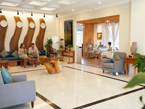 A hotel lobby with a reception desk, seating area with sofas and chairs, wooden wall decor, and a dining area with a table and chairs.