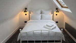 Una cama bien hecha con sábanas blancas y una toalla doblada, dos lámparas de noche, un tragaluz y un cabecero oscuro.