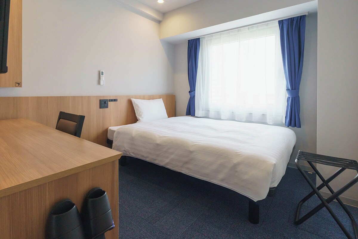 A hotel room with a bed, a desk, and a chair. There is a window with curtains.