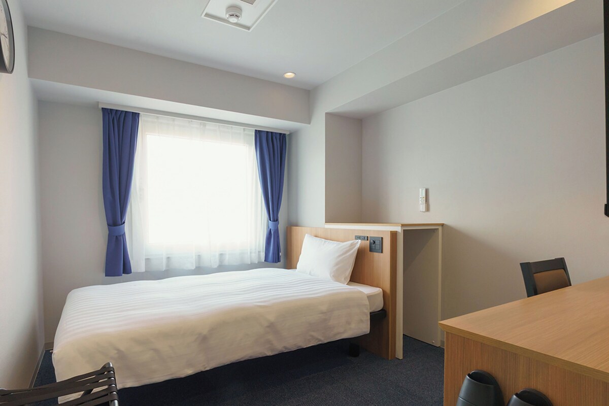 A hotel room with a bed, a desk, a chair, and a window with curtains.