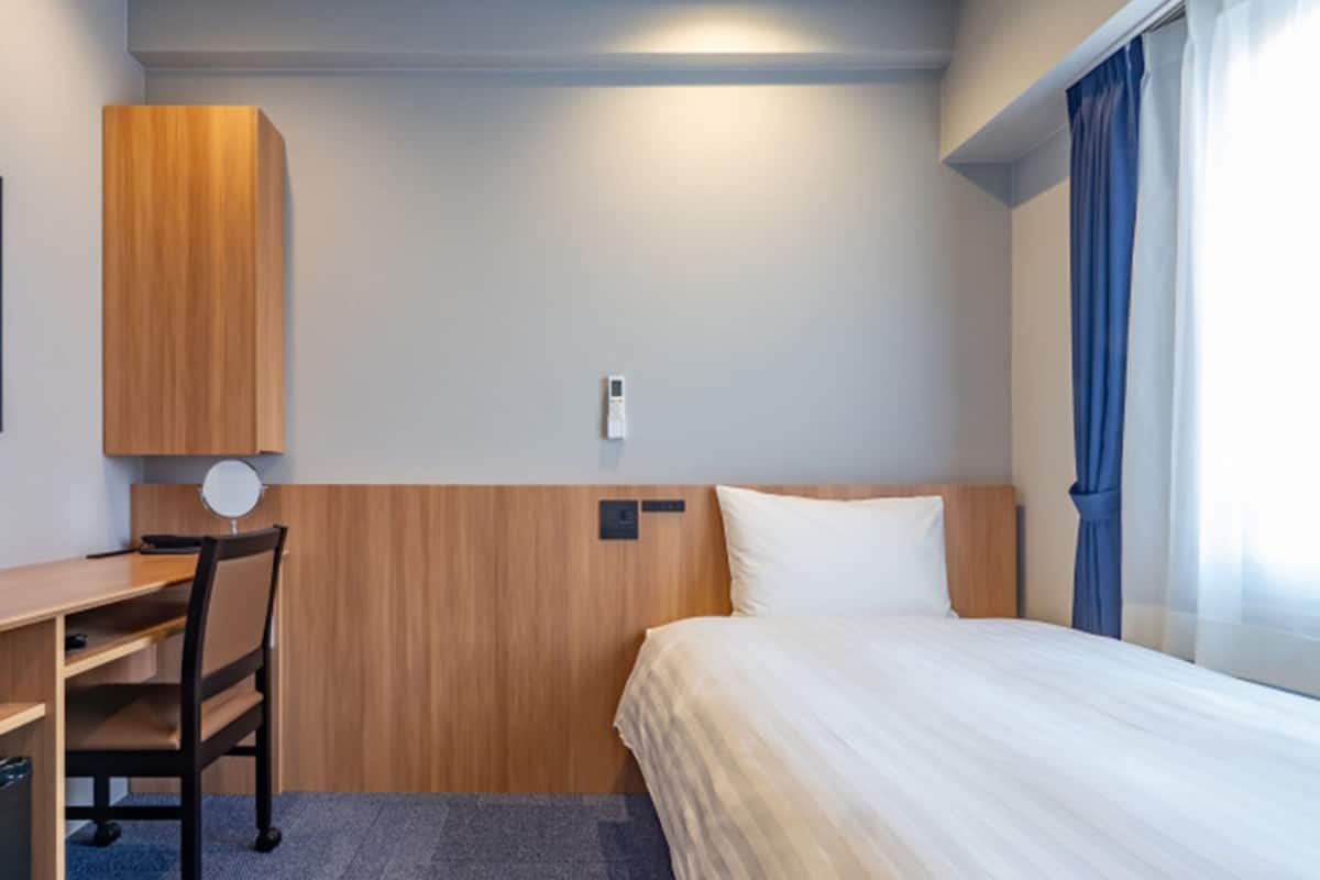 Quarto de hotel com cama, escrivaninha, cadeira, luminária na parede e janela com cortinas.