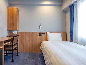 Quarto de hotel com cama, escrivaninha, cadeira, luminária na parede e janela com cortinas.