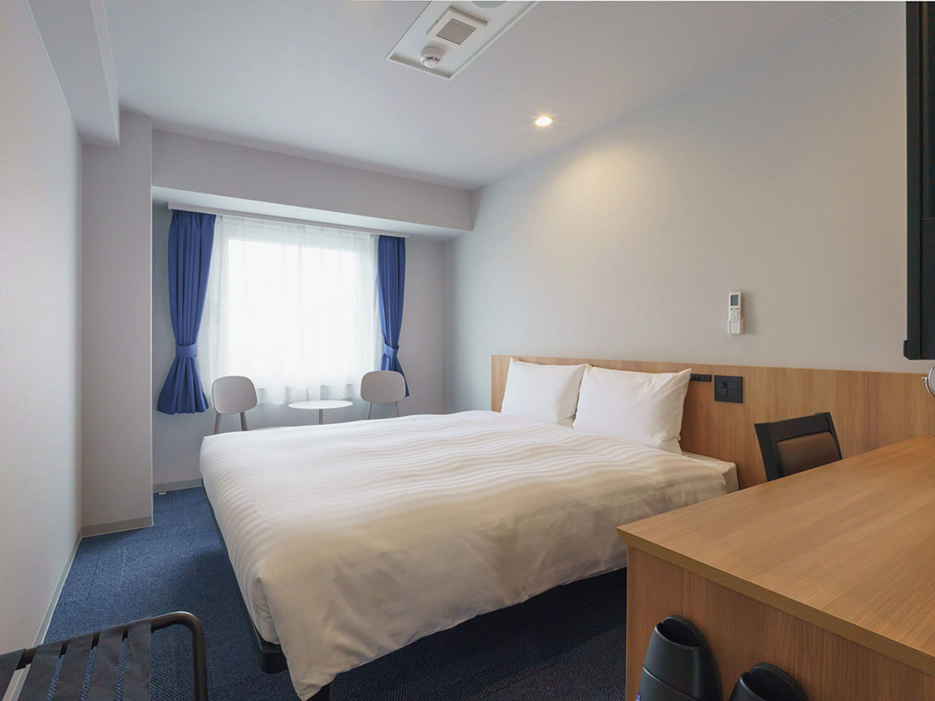 A hotel room with a large bed, a desk, two chairs, a window with curtains, and a ceiling light.