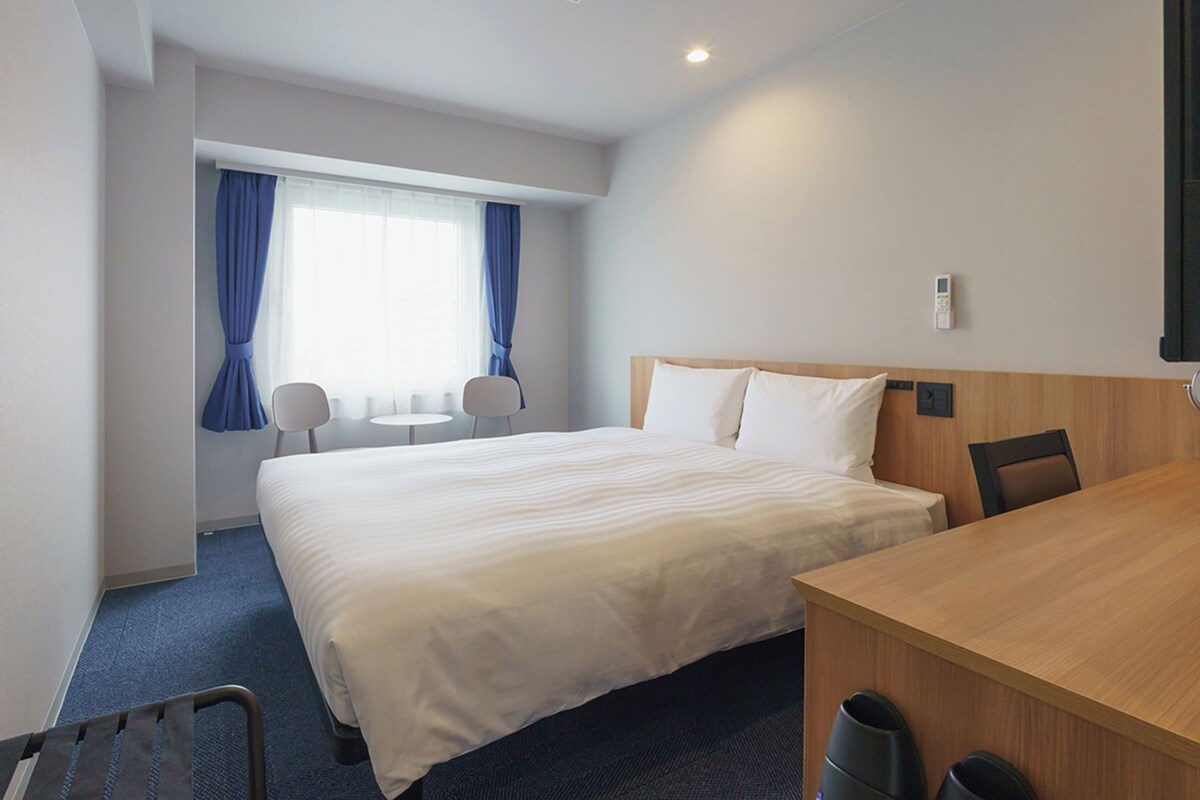 A hotel room with a large bed, a desk, two chairs, a window with curtains, and a ceiling light.
