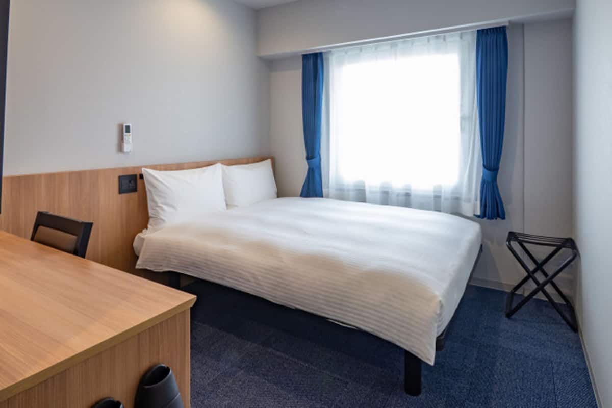 Quarto de hotel com cama, escrivaninha, cadeira, lixeira e janela com cortinas.
