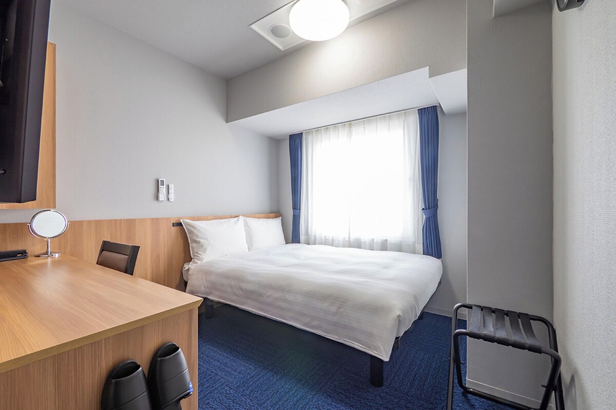 A hotel room with a bed, a desk, a chair, a mirror, and a window with curtains.