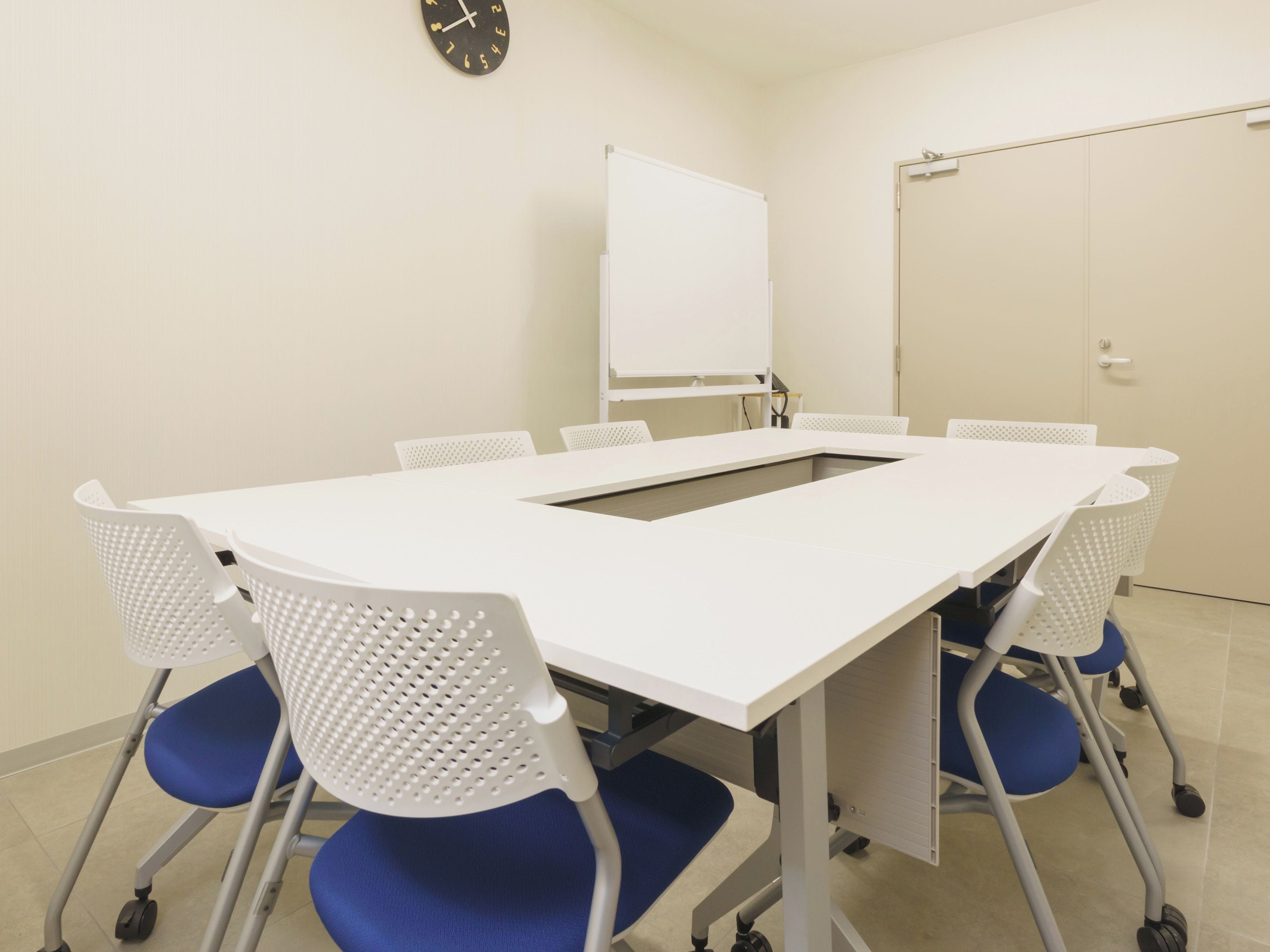 Une salle de conférence avec une table blanche, des chaises bleues, un tableau blanc et une horloge au mur.