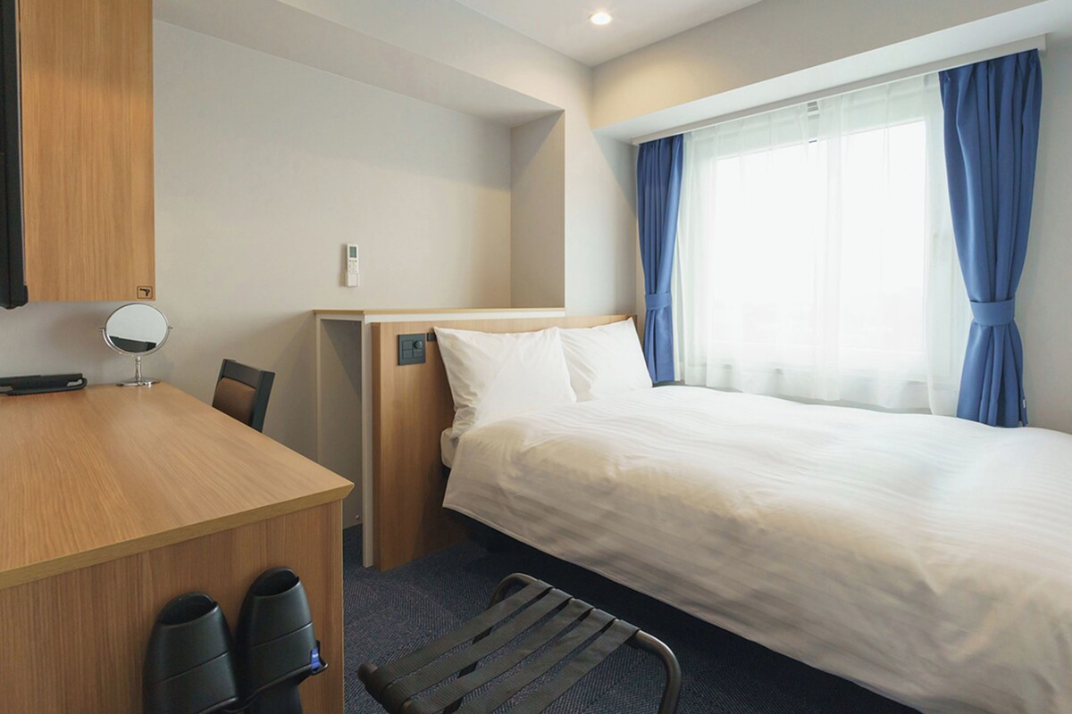 A hotel room with a bed, a desk, a chair, a mirror, and a window with curtains.