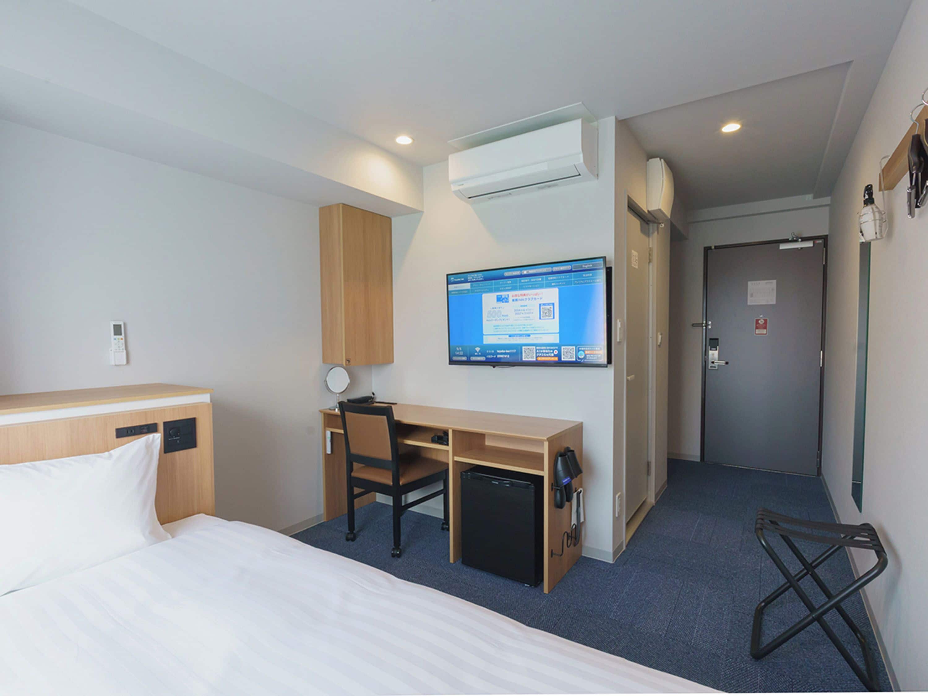 Une chambre d’hôtel comprenant un lit, un bureau avec une chaise, un petit réfrigérateur, un téléviseur à écran plat fixé au mur et une porte avec une poignée.