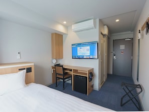 Habitación de hotel con cama, escritorio y silla, refrigerador pequeño, televisor de pantalla plana montado en la pared y puerta con manija.