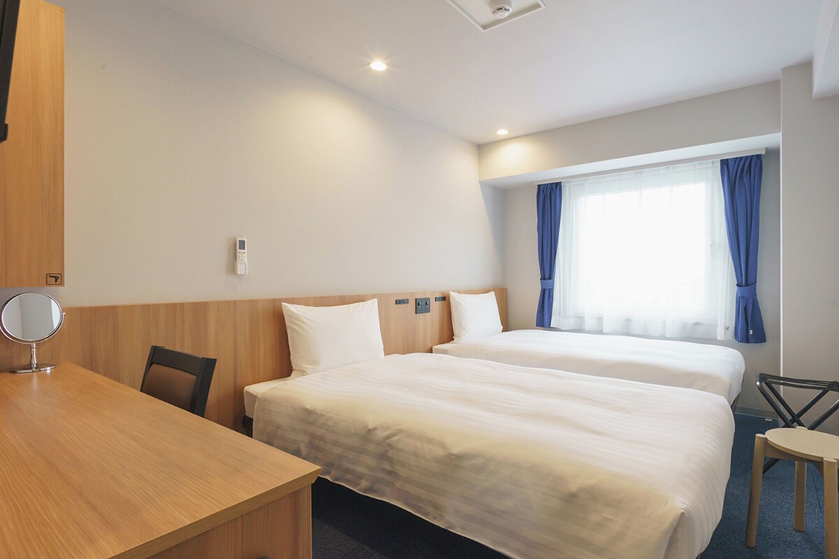 A hotel room with two beds, a desk, a chair, a small table, and a trash bin.