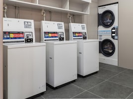 A row of three white washing machines with digital control panels, a wall-mounted washing machine, and a tiled floor.
