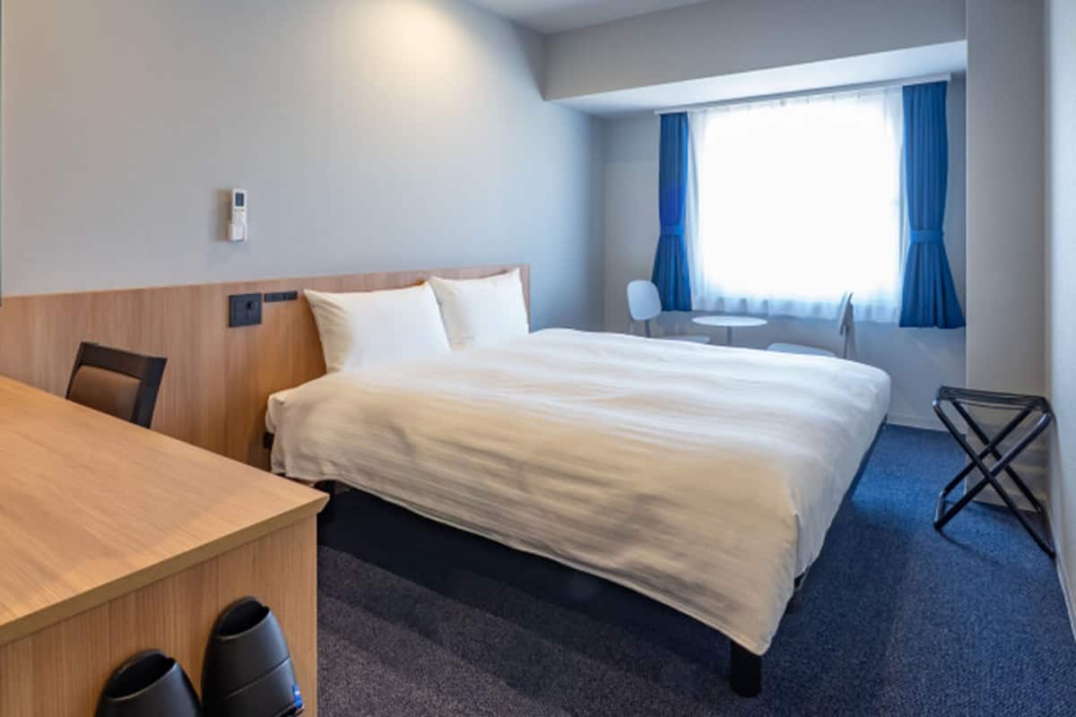 Quarto de hotel com cama, escrivaninha, cadeira, mesinha e janela com cortinas.