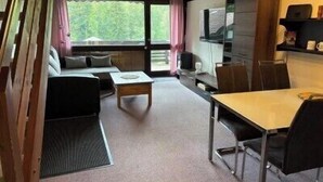 Una habitación con sofá, mesa de centro, mesa de comedor y sillas. Hay una ventana con cortinas, televisor y una estantería con objetos.