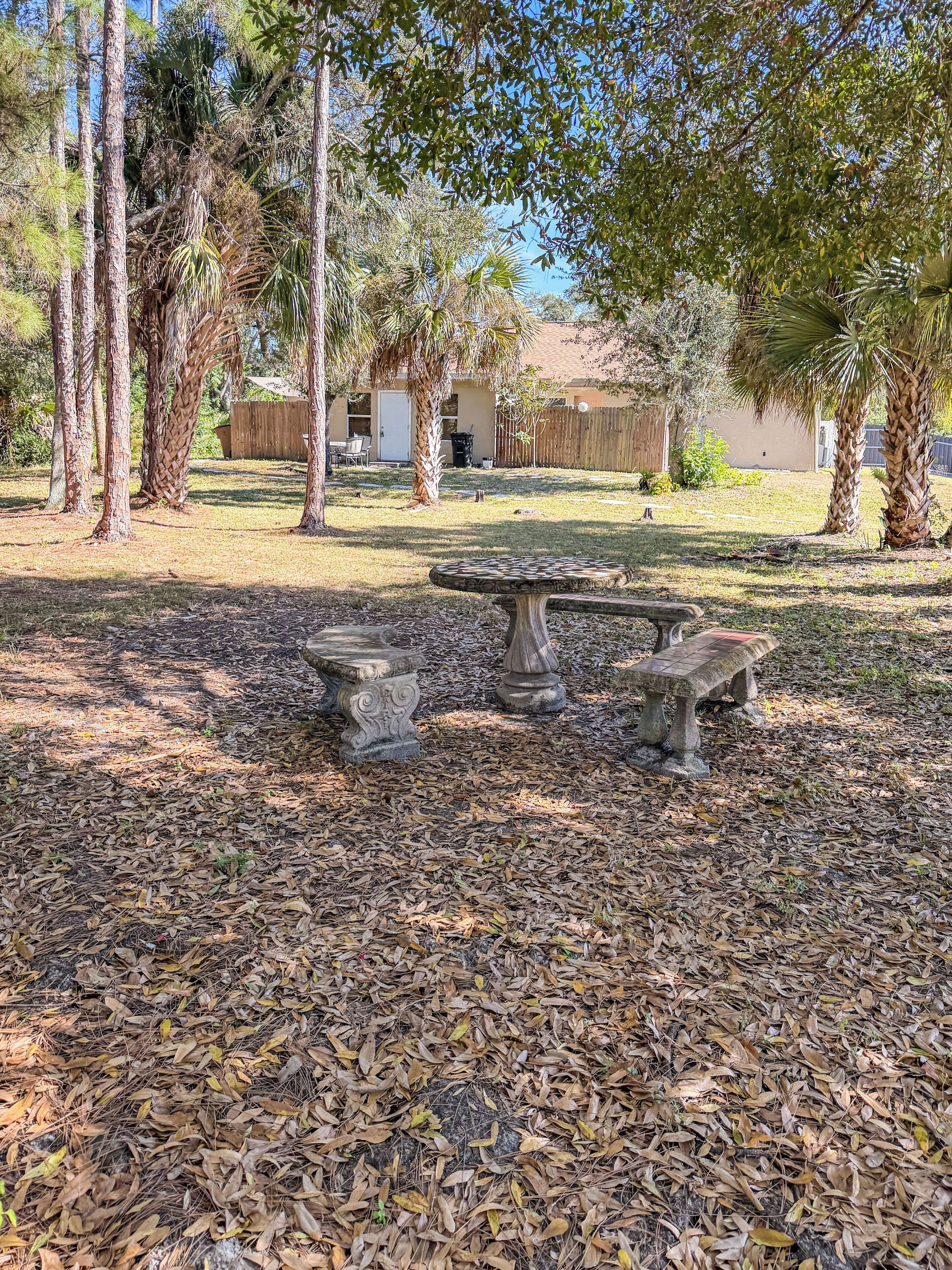 Une terrasse avec une table en pierre et des bancs, entourée d’arbres et d’une clôture en bois.