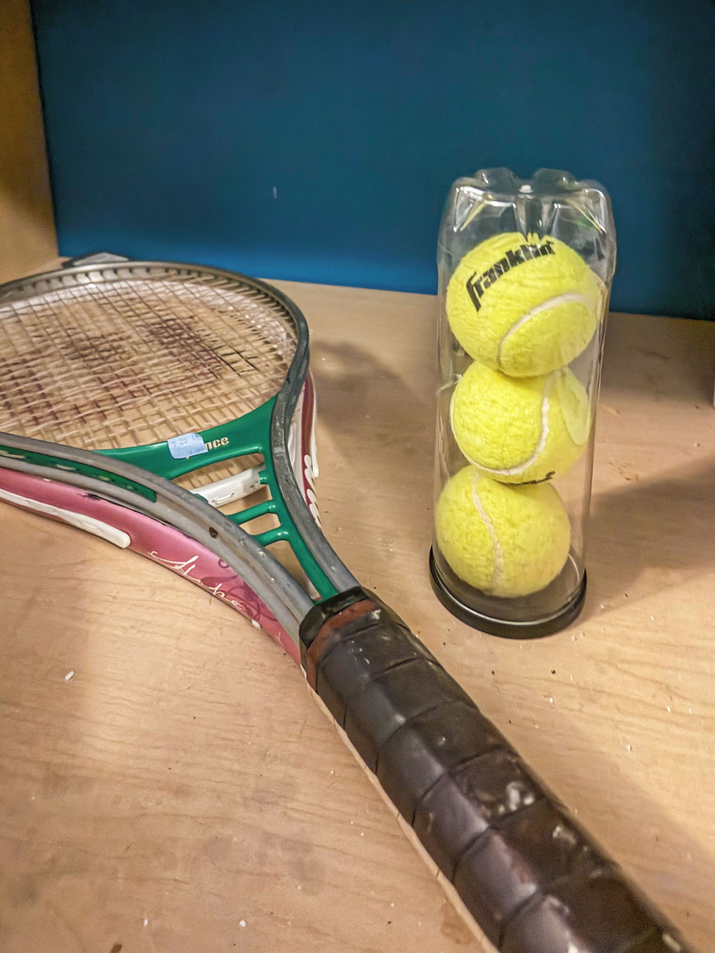 A tennis racket, a tennis ball container, and a tennis ball on a wooden surface.