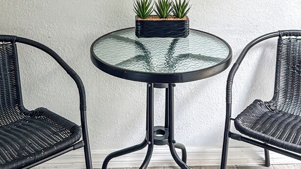 A round glass table with a black metal frame, two black wicker chairs, and a framed skull artwork on the wall. - Executive Studio (Lehigh Acres)