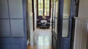 Een smalle gang met een deur naar een kamer met een raam, een kleine tafel met stoelen en een bloemenvaas.