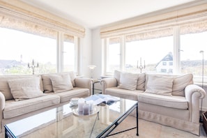 Ein geräumiges Wohnzimmer mit zwei beigen Sofas, einem Glastisch und großen Fenstern mit Vorhängen.