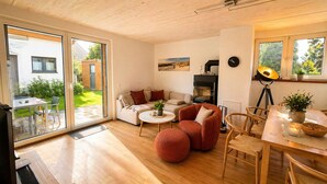 Ein modernes Wohnzimmer mit einer Couch, einem Couchtisch und einem roten Sessel, ein Essbereich mit Tisch und Stühlen, ein Kamin und große Fenster.