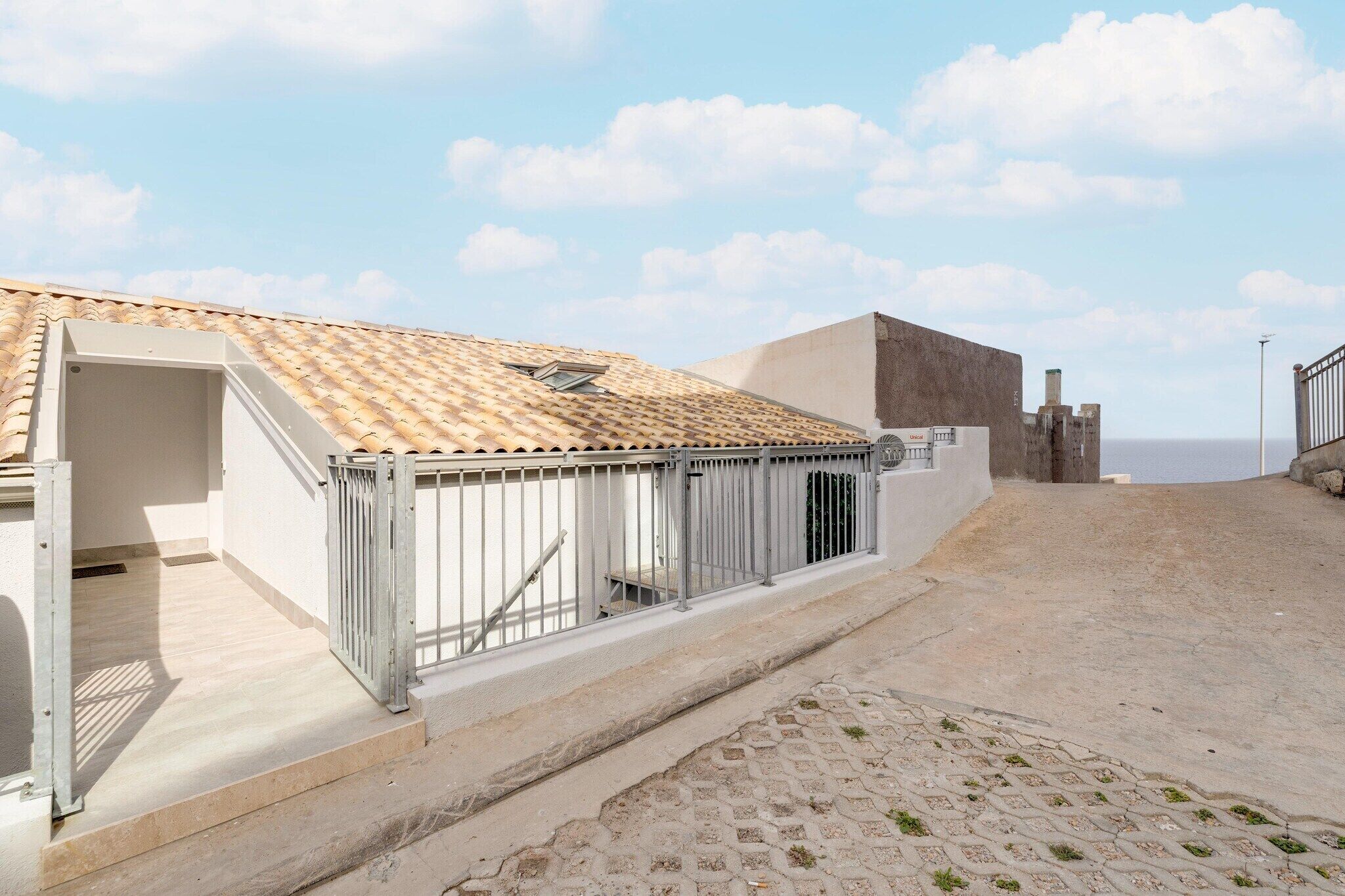 A building with a tiled roof, a metal fence, and a paved area leading to a gate.