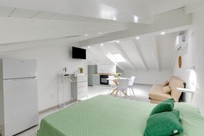 Ein modernes Schlafzimmer im Loft-Stil mit grüner Bettdecke, einer Küchenzeile, einem Essbereich und einem an der Wand montierten Fernseher.