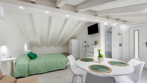 A modern, well-lit room with a bed, a dining table with chairs, a TV mounted on the wall, and a kitchenette area. - Attic studio, with air conditioning (Castelsardo)