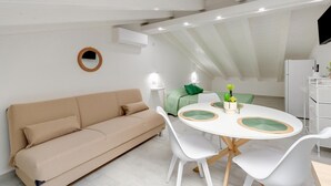 A modern, well-lit room with a white sofa, a round dining table with white chairs, and a small kitchenette area. - Attic studio, with air conditioning (Castelsardo)