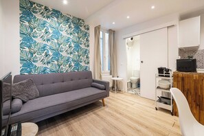 A modern living room with a grey sofa, a small round table, a white chair, a wooden sideboard, and a kitchen area with a white cabinet and a black appliance. - Cozy Apartment a/c 1BR/2P - Nice (Nice)
