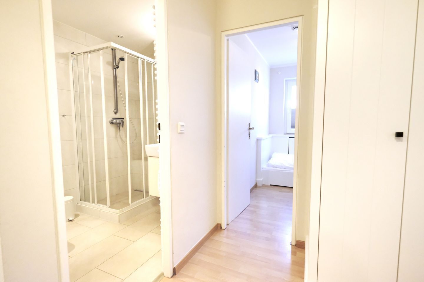 En gang der fører til et badeværelse med et brusekabine i glas, et toilet og en seng i det tilstødende rum.