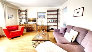 Uma sala de estar com um sofá vermelho, um sofá roxo, uma mesa de centro com revistas, uma televisão e uma estante de livros.