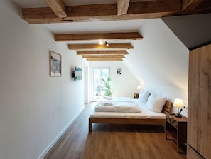 Une chambre avec un plafond en pente, des poutres en bois, un lit avec une literie blanche, une table de chevet avec une lampe et une fenêtre donnant sur un paysage verdoyant.