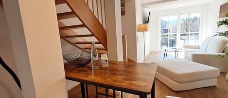 A modern kitchen with a wooden staircase, a dining table with stools, and a view of the outdoors through large windows.