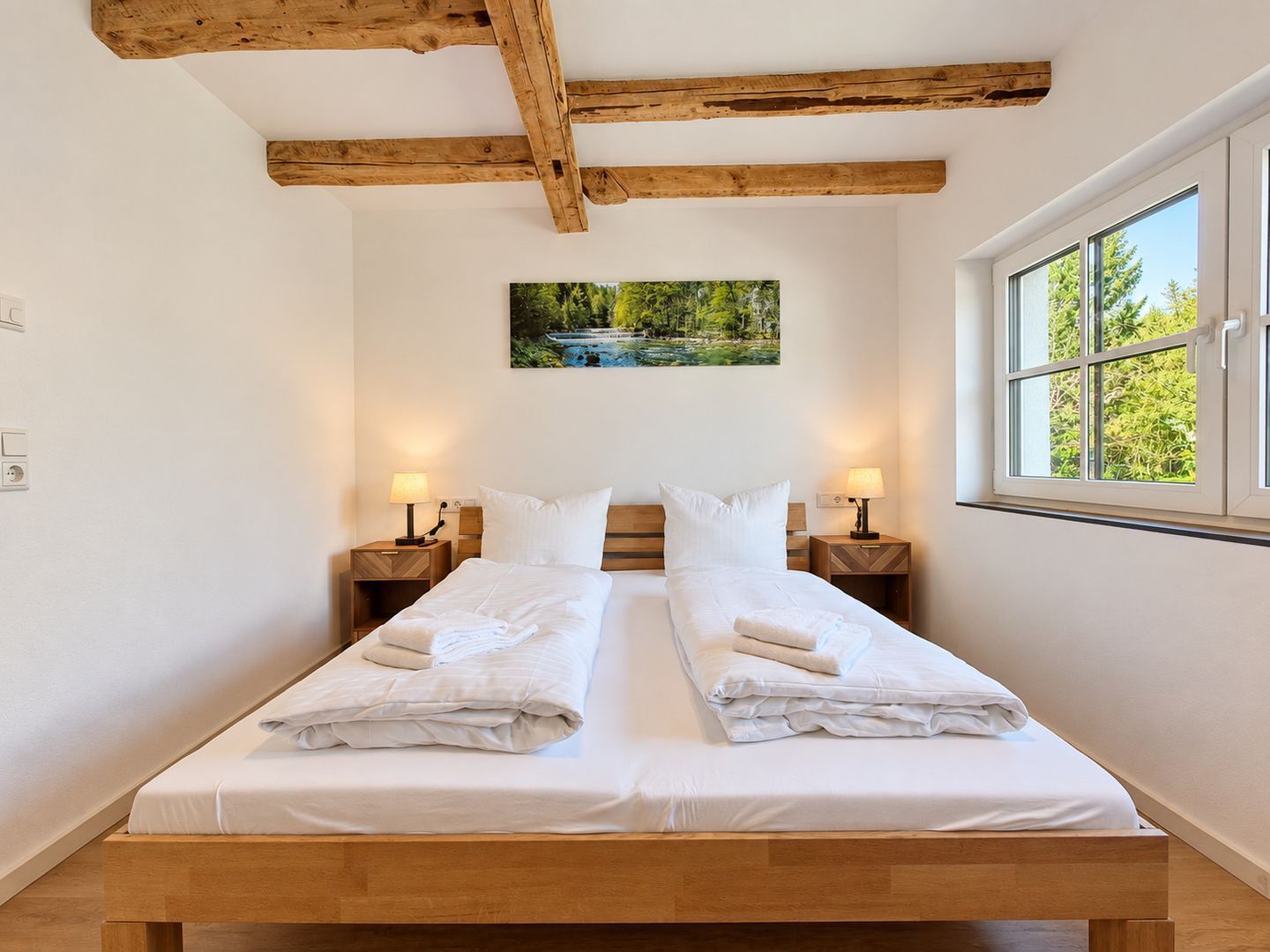 A double bed with white linens, wooden headboard, two bedside tables with lamps, a painting of a landscape, and a window with a view of trees.