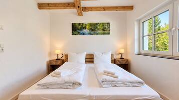 A double bed with white linens, wooden headboard, two bedside tables with lamps, a painting of a landscape, and a window with a view of trees.