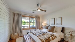 Un dormitorio con cama, una banca, ventilador de techo, ventana con cortinas y cuadros en la pared.